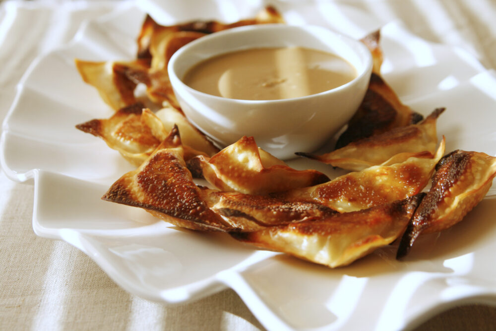 baked peanut chicken wontons are shown on a white plate with a bowl of peanut dipping sauce. The image has  sunlight lines across it from blinds.