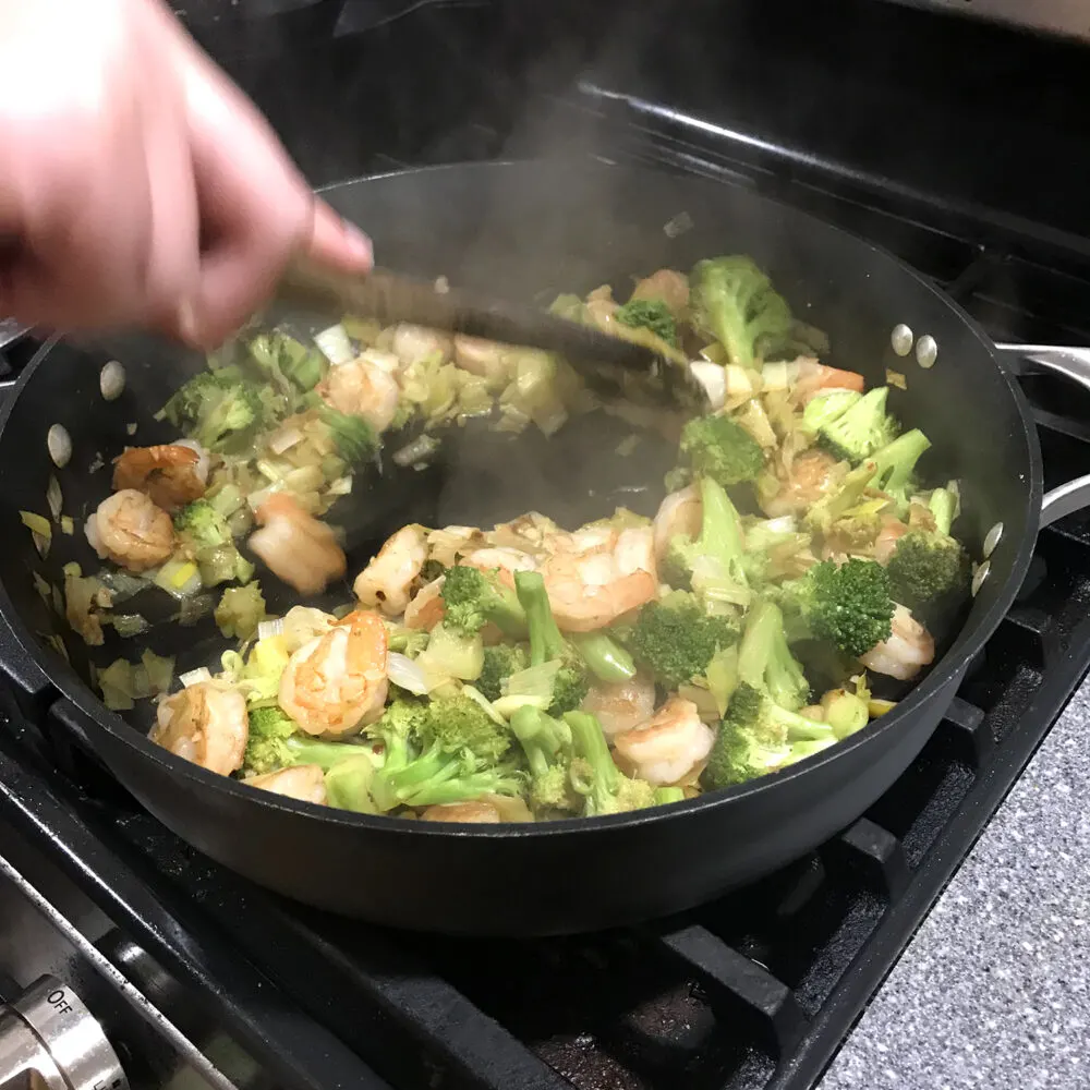 A hand holds a wooden spatula, stirring inside a large skillet on the stove.