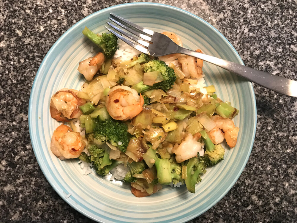 Easy Garlicky Leek, Broccoli and Shrimp Stir-Fry is shown in a blue bowl with a silver fork on a granite countertop.