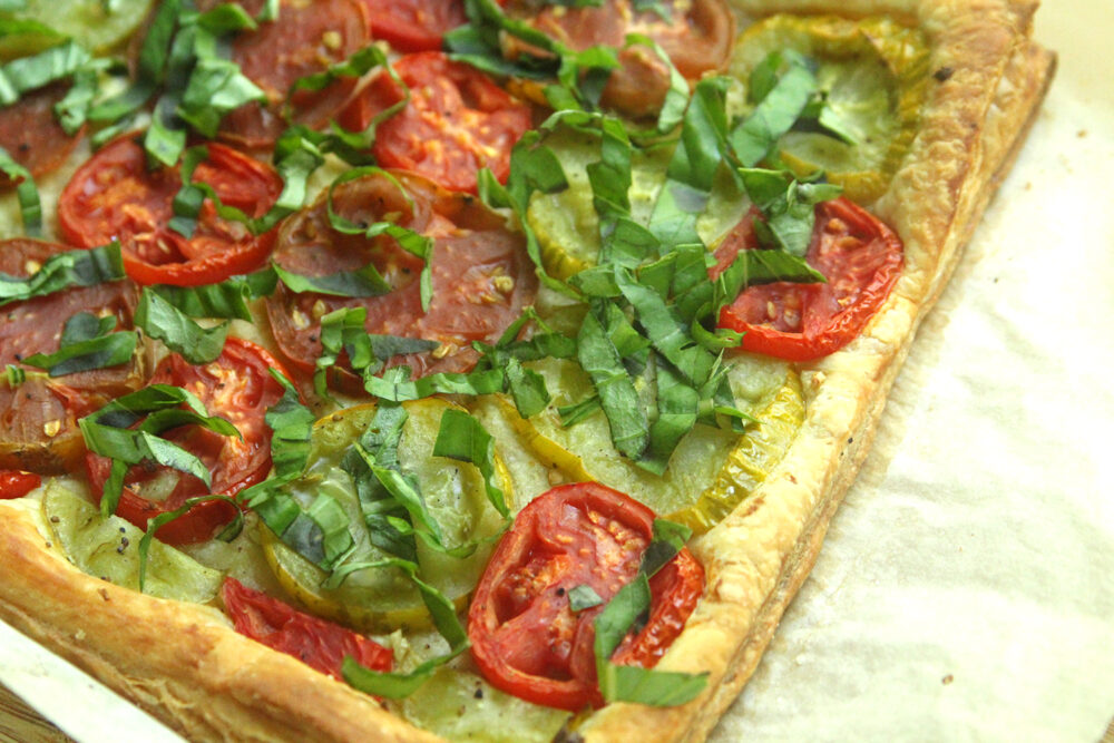 A browned puff pastry topped with red and green tomatoes and fresh basil sits on a sheet of parchment pepper.