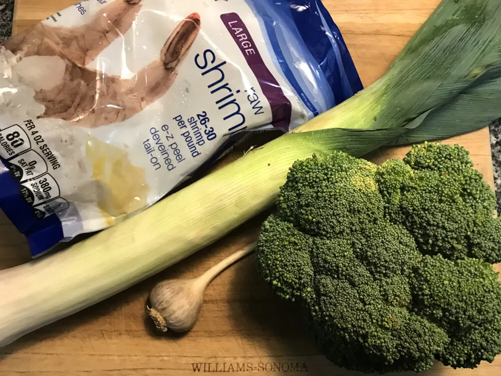 Ingredients are shown on a wooden cutting board: frozen shrimp, a leek, garlic and broccoli.