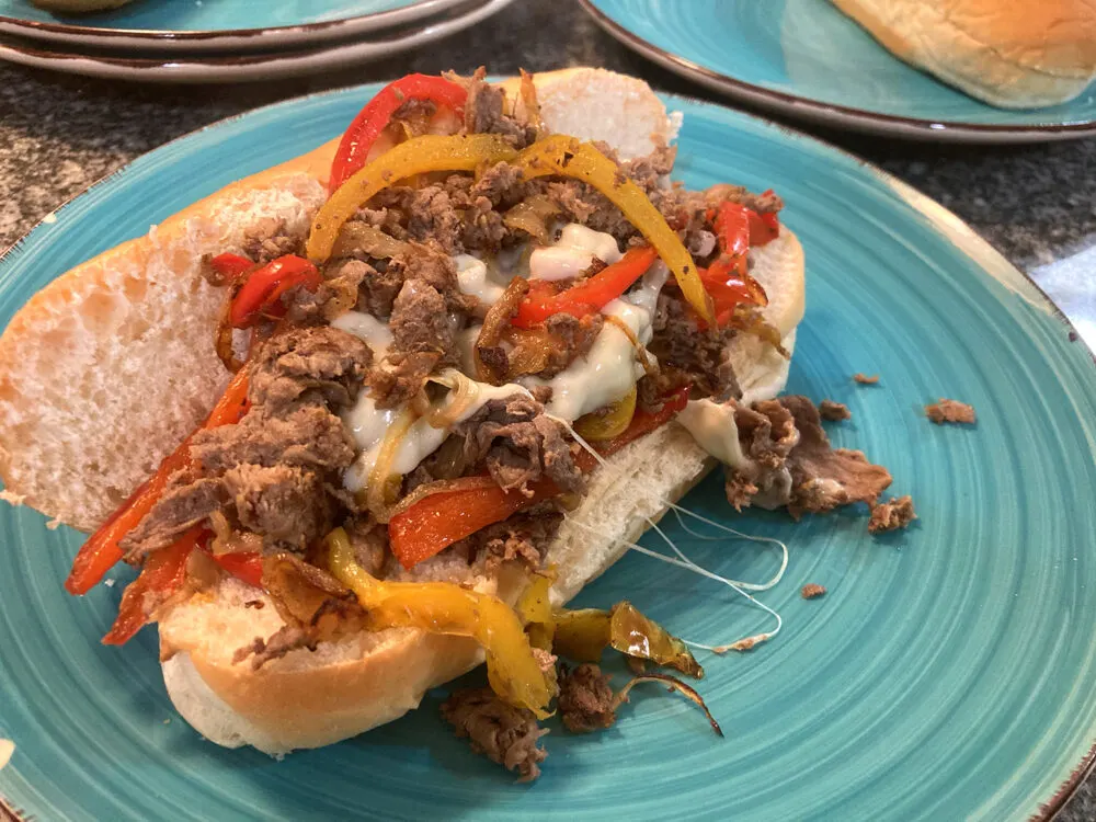 A long submarine roll holds sauteed steak, peppers, onions and cheese on a turquoise plate.