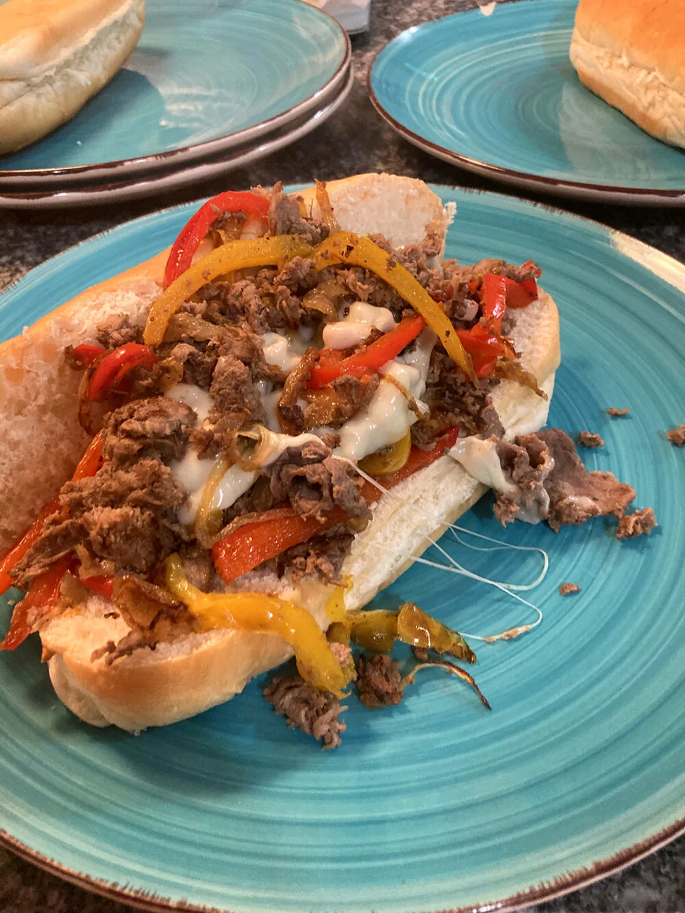 A long submarine roll holds sauteed steak, peppers, onions and cheese on a turquoise plate.