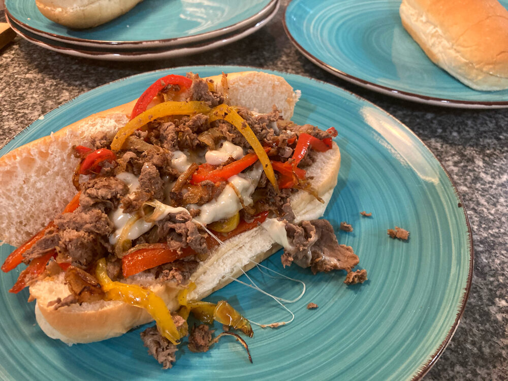 A long submarine roll holds sauteed steak, peppers, onions and cheese on a turquoise plate.
