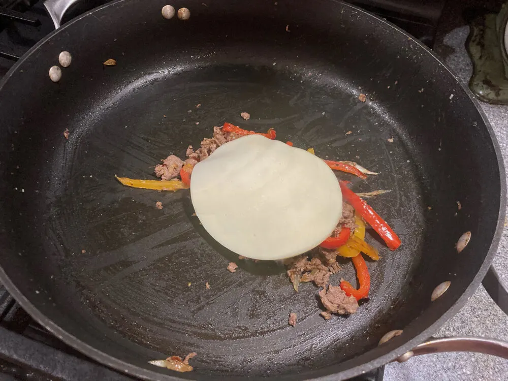 A round slice of ivory-colored cheese sits on steak, peppers and onions in a skillet.