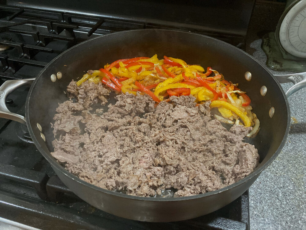 Peppers, onions and shaved steak are shown sauteing in a large skillet. Steam is rising from the skillet.