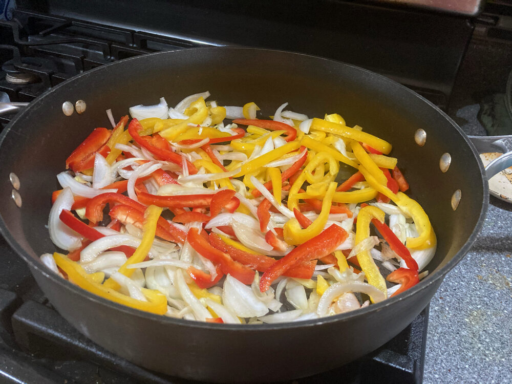 Red peppers, yellow peppers and onions are shown sauteing in a large skillet.