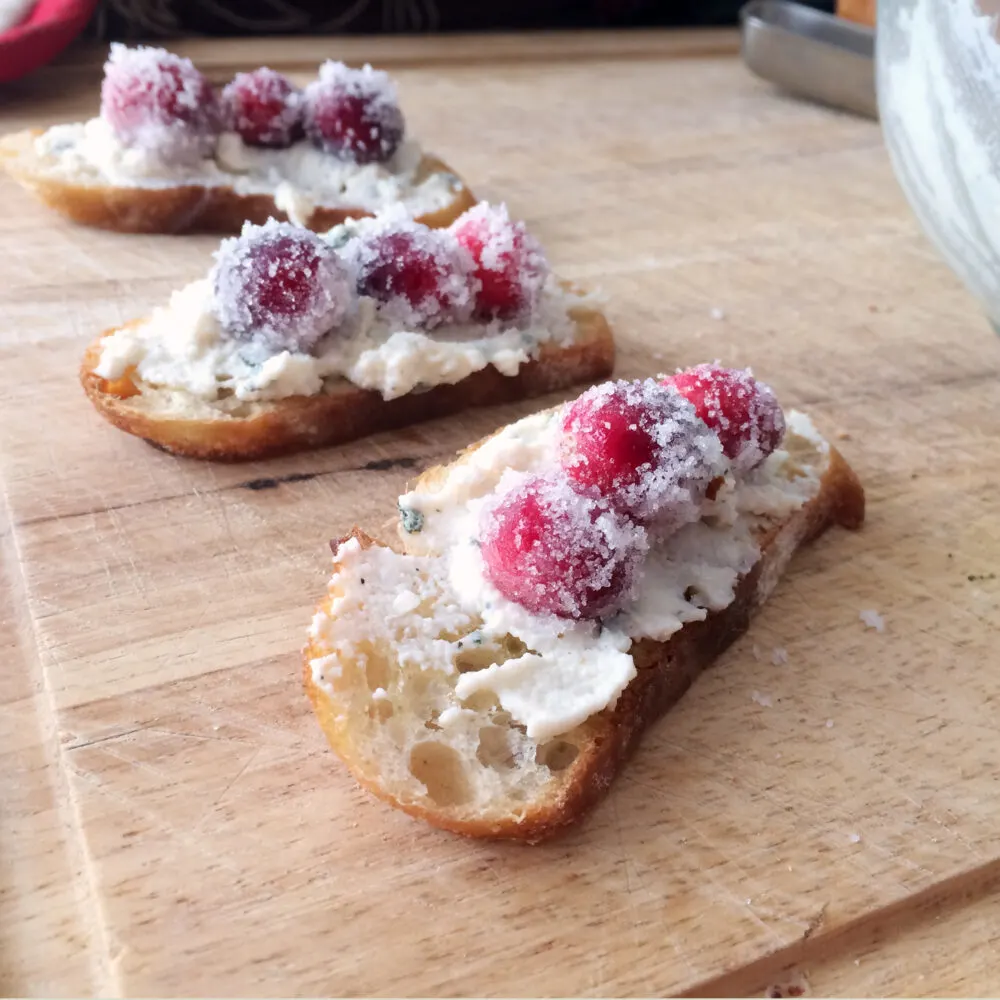 Three crostini topped with sage ricotta and sugared cranberries sit on a cutting board.