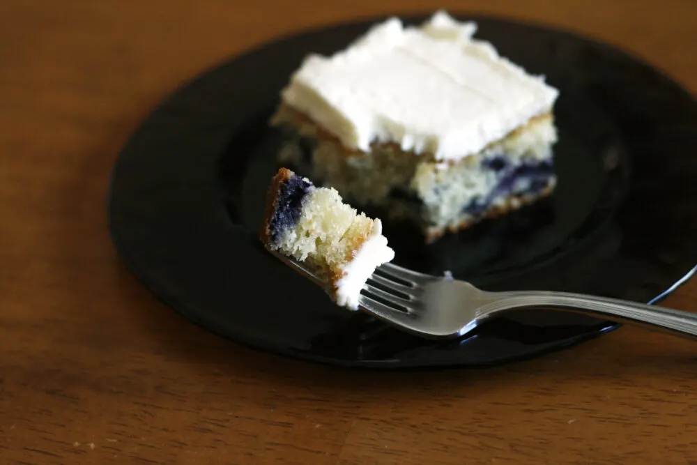 A bite of Blueberry Cake with Fluffy Vanilla Frosting is on a fork. Nearby the slice of cake sits on a purple glass plate on a wooden table.