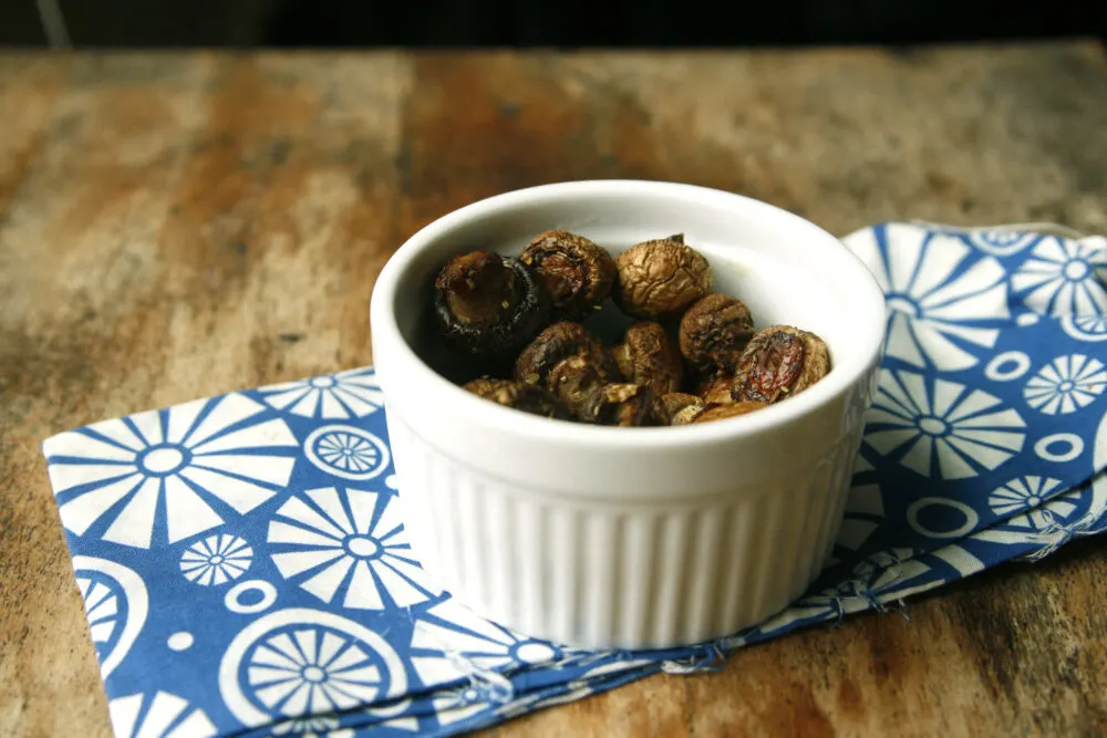 A white bowl of Herbed Roasted Mushrooms sits on a blue and white napkin on a wood table.