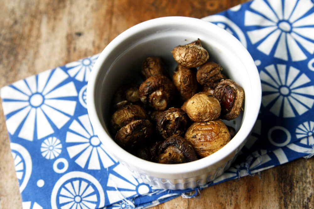 A white bowl of browned Herbed Roasted Mushrooms sits on a blue and white napkin on a wooden table.