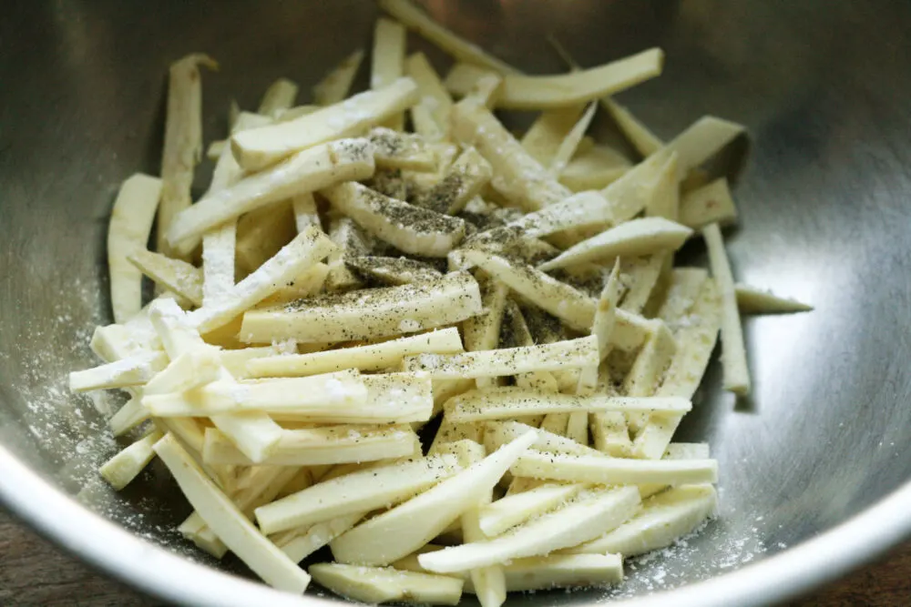 Peeled parsnips cut into fry-like shapes are shown in a silver bowl with seasonings and cornstarch.