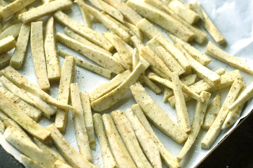 Uncooked Baked Peppery Parsnip Fries are shown on a parchment-lined baking sheet. 