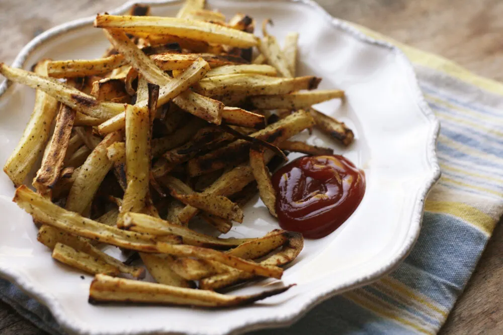 a plate of Baked Peppery Parsnip Fries with ketchup is shown on a blue and yellow napkin.