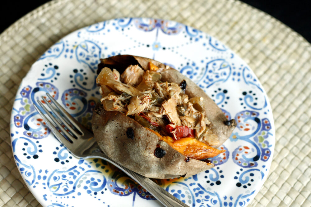 An overhead shot of a baked sweet potato stuffed with pulled chicken and red peppers on a colorful plate on a tan woven mat on a black table.
