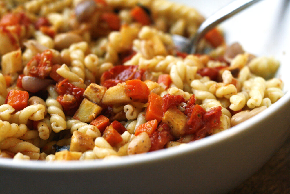 Roasted Vegetable Pasta with Tomatoes and White Beans is shown in a white bowl with a silver-toned serving spoon.