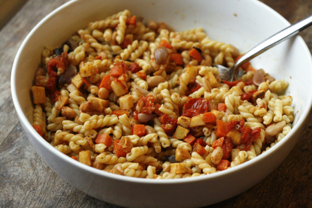 Roasted Vegetable Pasta with Tomatoes and White Beans is held in a white bowl with a serving spoon in it. The bowl is on a wooden surface.