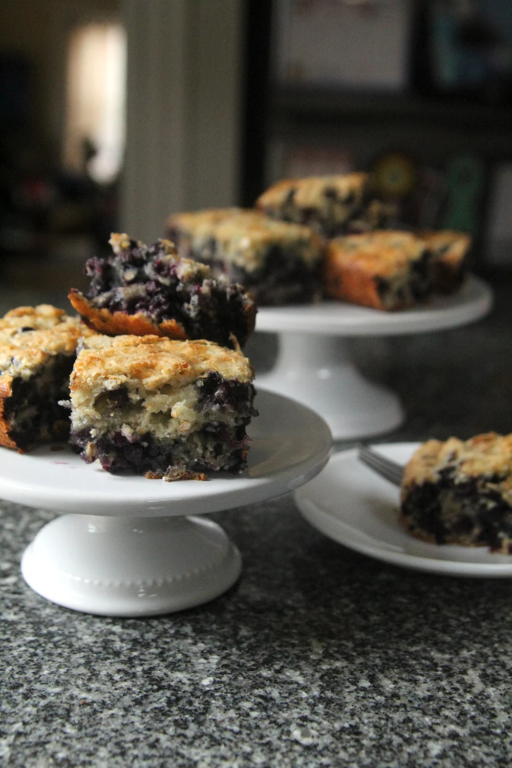 Piles of Blueberry Oatmeal Cake sit on two white cake plates. A small white plate holds another piece with a silver fork.