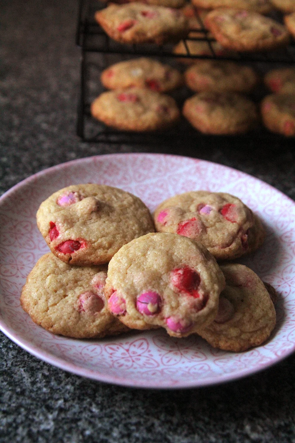 A pink and white plate on a granite countertop shows a pile of M&M cookies for Valentine's Day dotted with red and pink candies. A cooling rack with cookies sits in the background.