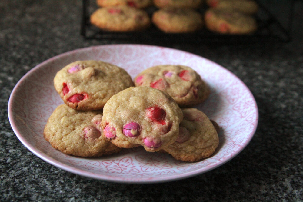 A pink and white plate on a granite countertop shows a pile of cookies for Valentine's Day dotted with red and pink candies. Nearby, a cooling rack with cookies sits.