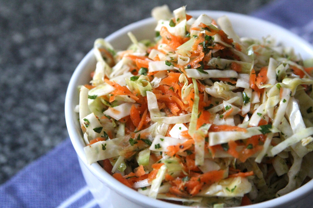 Easy Oil and Vinegar Coleslaw is shown in a white bowl on a blue towel on a gray granite countertop.