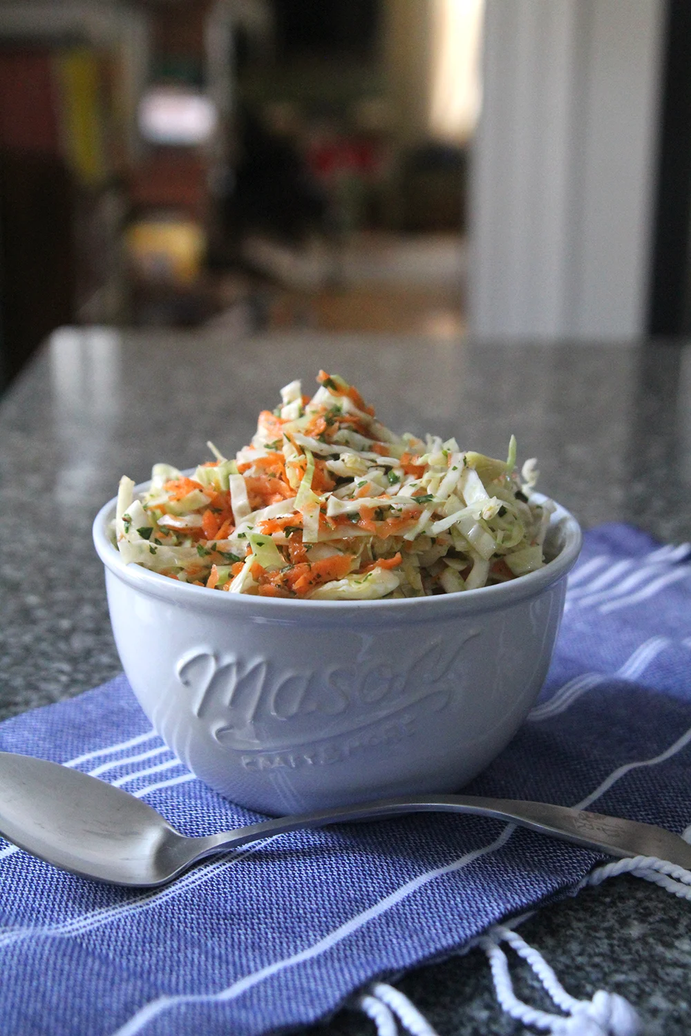 Oil and Vinegar Coleslaw is shown in a white bowl on a blue and white cloth on a granite countertop with a silver spoon nearby.