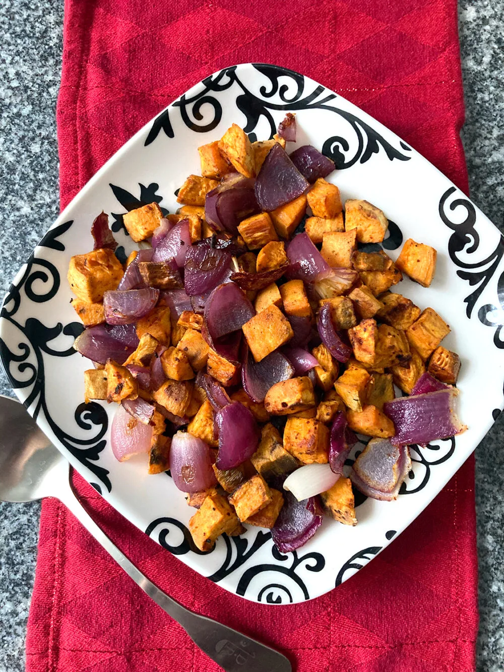 An overhead shot of Roasted Sweet Potatoes and Red Onions with Gochujang Seasoning recipe on a black and white plate on a red napkin on a granite countertop.