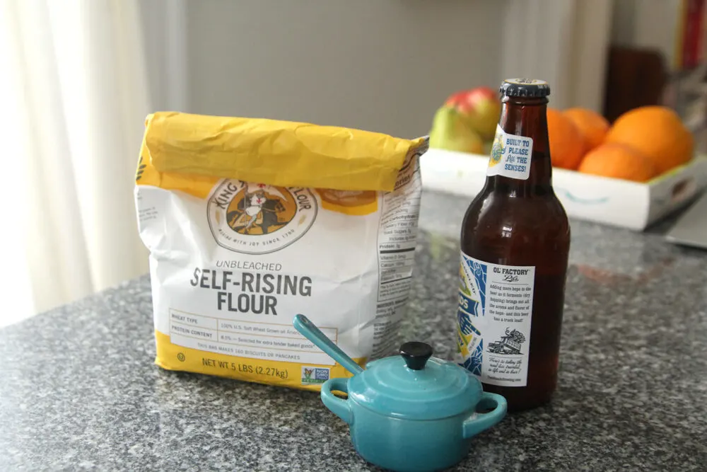 Easy Beer Bread is made with three ingredients pictured here: self-rising flour, beer and salt. These are shown on a granite countertop with a tray of fruit in the background.