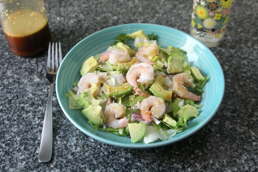 A salad featuring shrimp, avocado and red onions dressed with Sesame Ginger Dressing sits on a granite countertop with a fork, more dressing and a glass of water nearby.