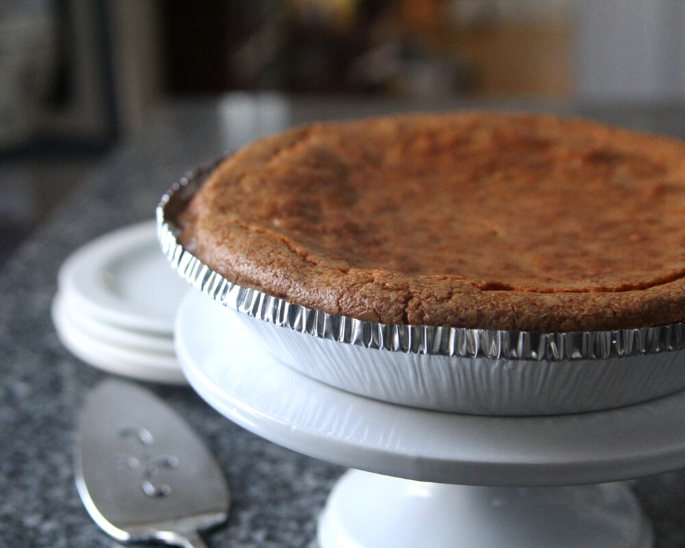 Chocolate Chip Pie is shown in a silver pie plate on a white pie stand on a gray granite countertop. There is a stack of white plates nearby and a silver pie knife.