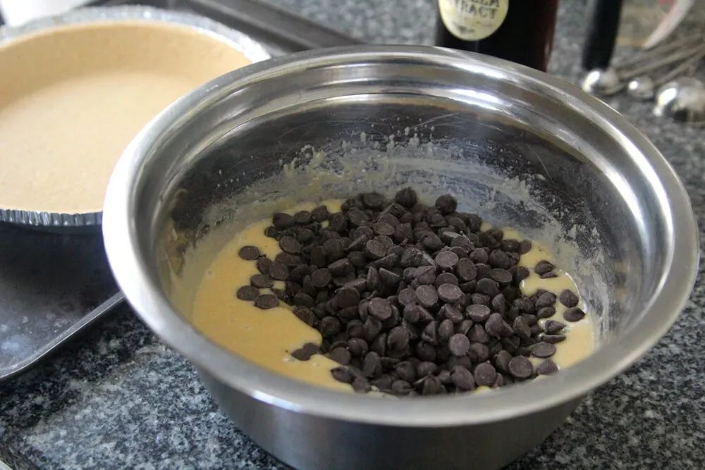 The batter for Chocolate Chip Pie is shown in a silver bowl. A bottle of vanilla extract, measuring spoons and a pie shell are shown nearby.