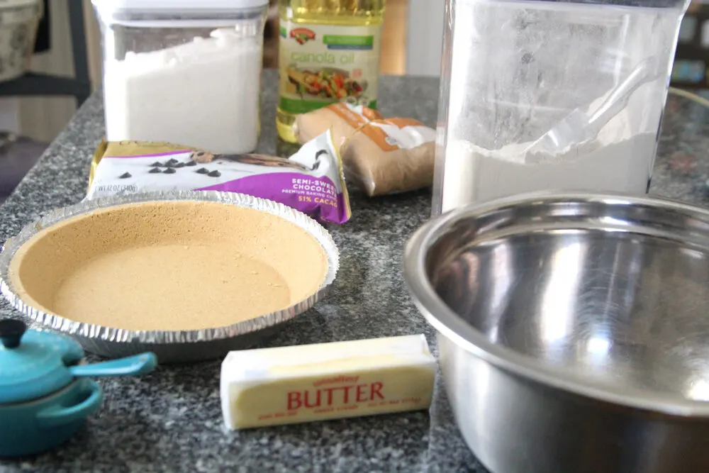 Ingredients for Chocolate Chip Pie are shown on a granite countertop including butter, a pie shell, chocolate chips, white sugar, brown sugar, canola oil and flour.