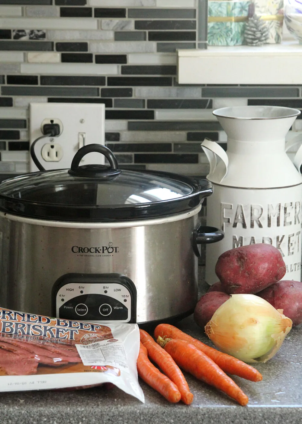 A slow cooker, potatoes, carrots, an onion and a package of corned beef sit on a counter.