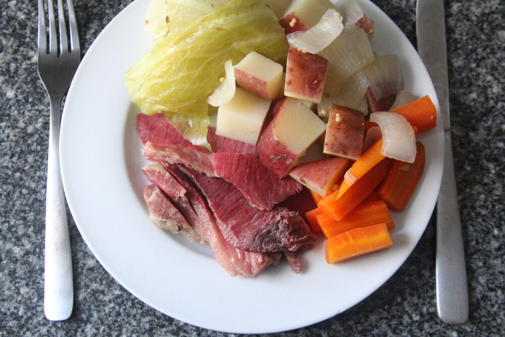 A white plate with a fork and knife next to it sits on a granite countertop. On the plate is corned beef, cabbage, carrots, potatoes and onions.