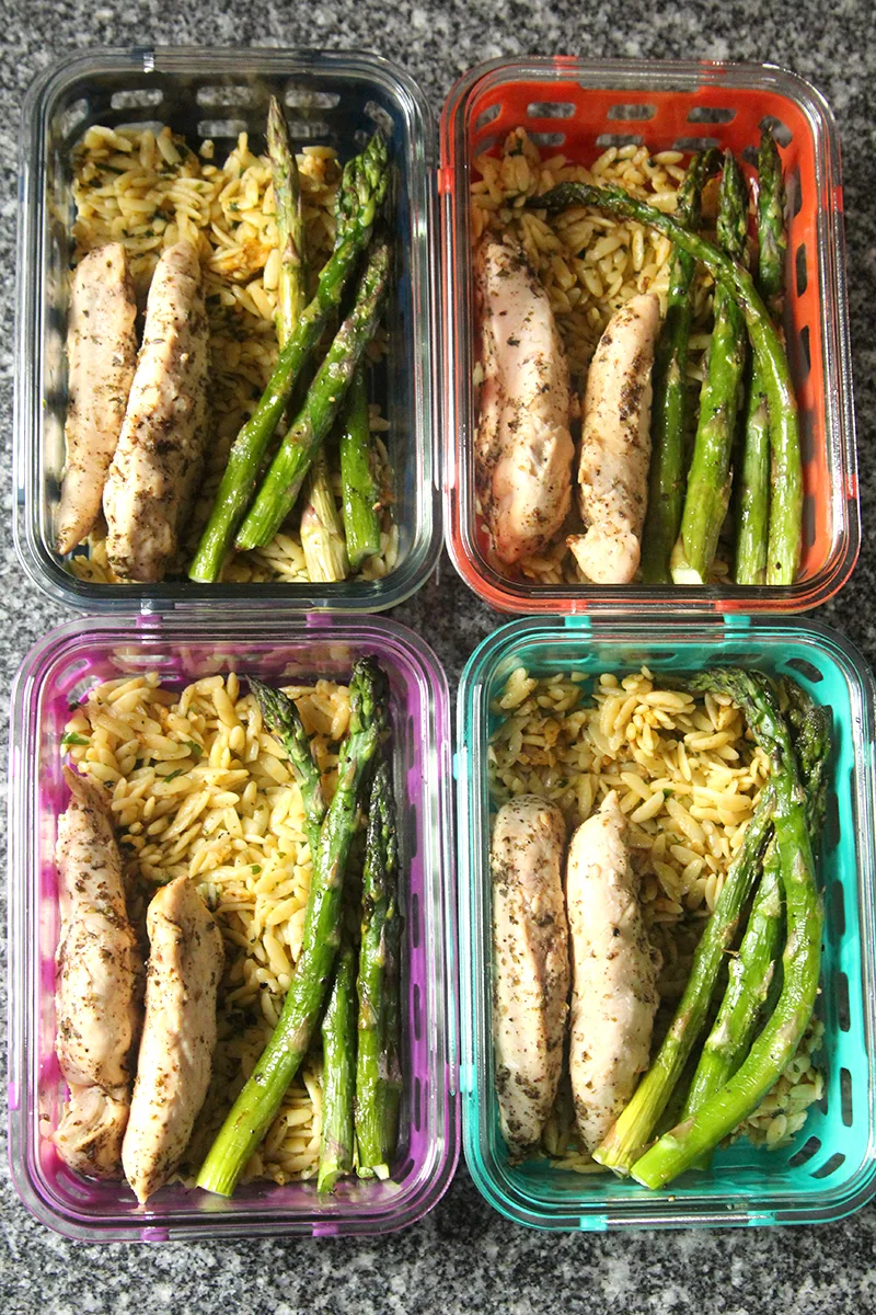 Four silicone-covered, glass meal prep containers are shown from above on a countertop. Each contains seasoned orzo, cooked chicken with herbs and asparagus.