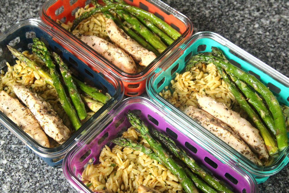 Four silicone-covered, glass meal prep containers are shown on a countertop. Each contains seasoned orzo, cooked chicken with herbs and asparagus.