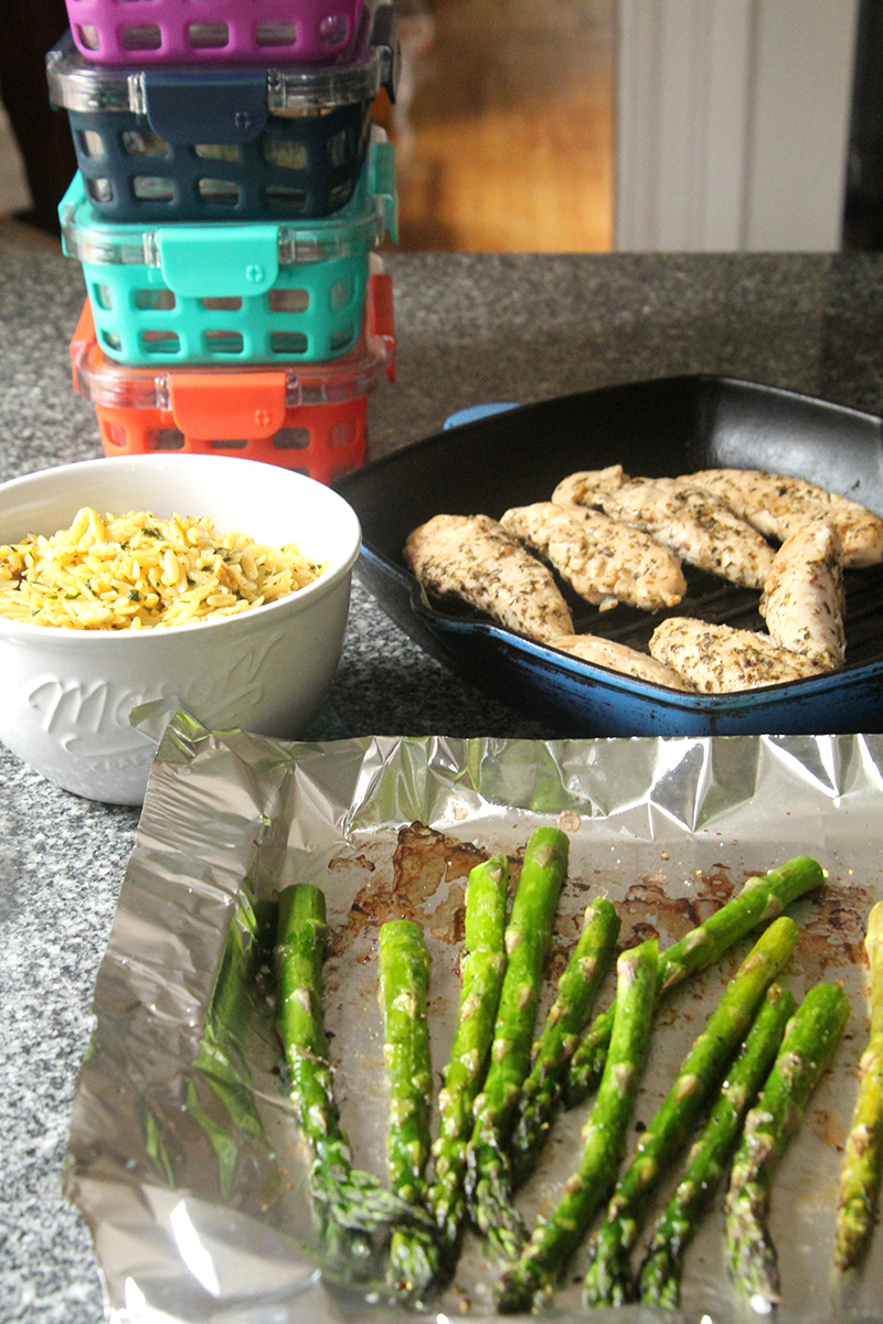 On a kitchen counter sits a bowl of seasoned orzo, a grill pan with herb-covered chicken, a foil-lined sheet pan with roasted asparagus and four meal prep containers.