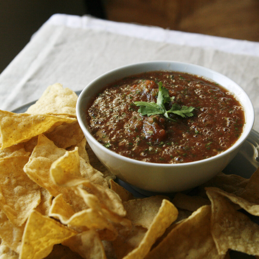 A white bowl filled with red salsa specked with green and topped with cilantro leaves sits surrounded by tortilla chips on a plate.