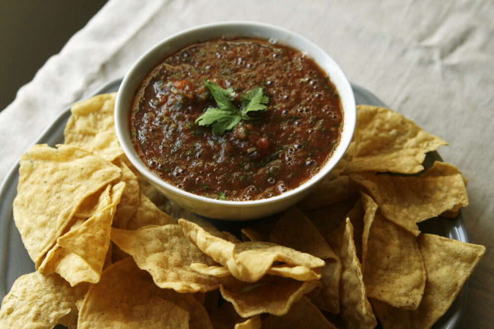 A white bowl filled with red Spicy Restaurant Style Salsa specked with green and topped with cilantro leaves sits surrounded by tortilla chips on a plate. It's situated on a linen cloth.