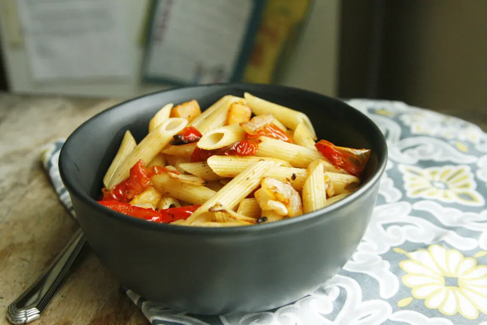 A bowl of Roasted Vegetable Pasta Toss featuring red peppers and penne pasta sits on a grey, yellow and white mat on a wooden table with a fork nearby.