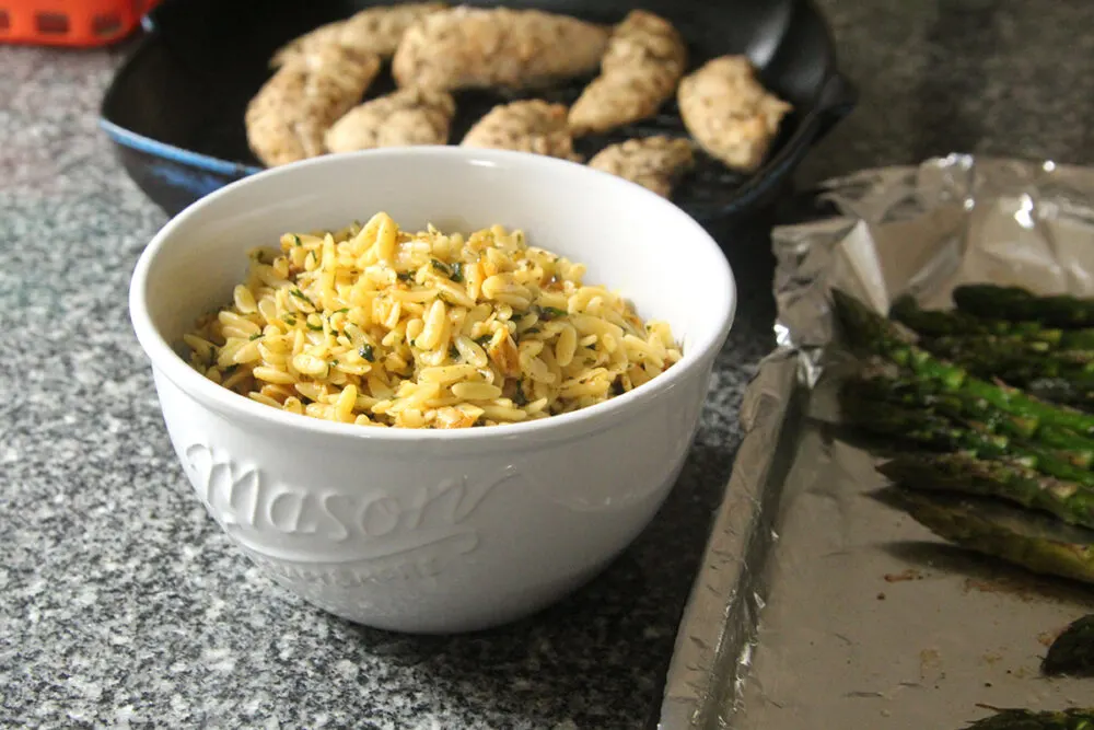 A bowl of lemon butter orzo sits next to a tray of roasted asparagus and a pan of chicken on a gray granite countertop.