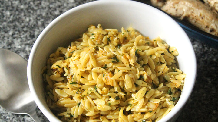 A bowl of lemon butter orzo sits on a counter next to a pan of chicken and a silver serving spoon.