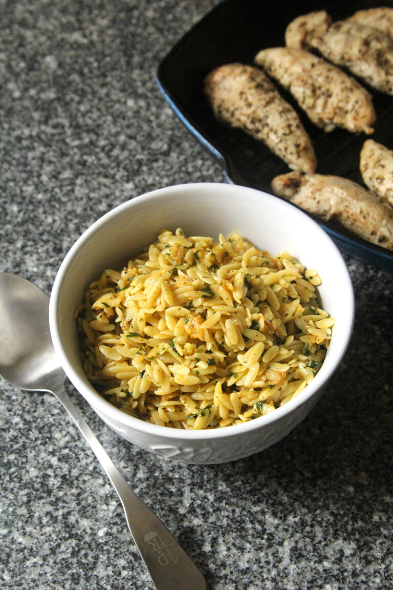 A bowl of lemon butter orzo sits on a counter next to a pan of chicken and a silver serving spoon.