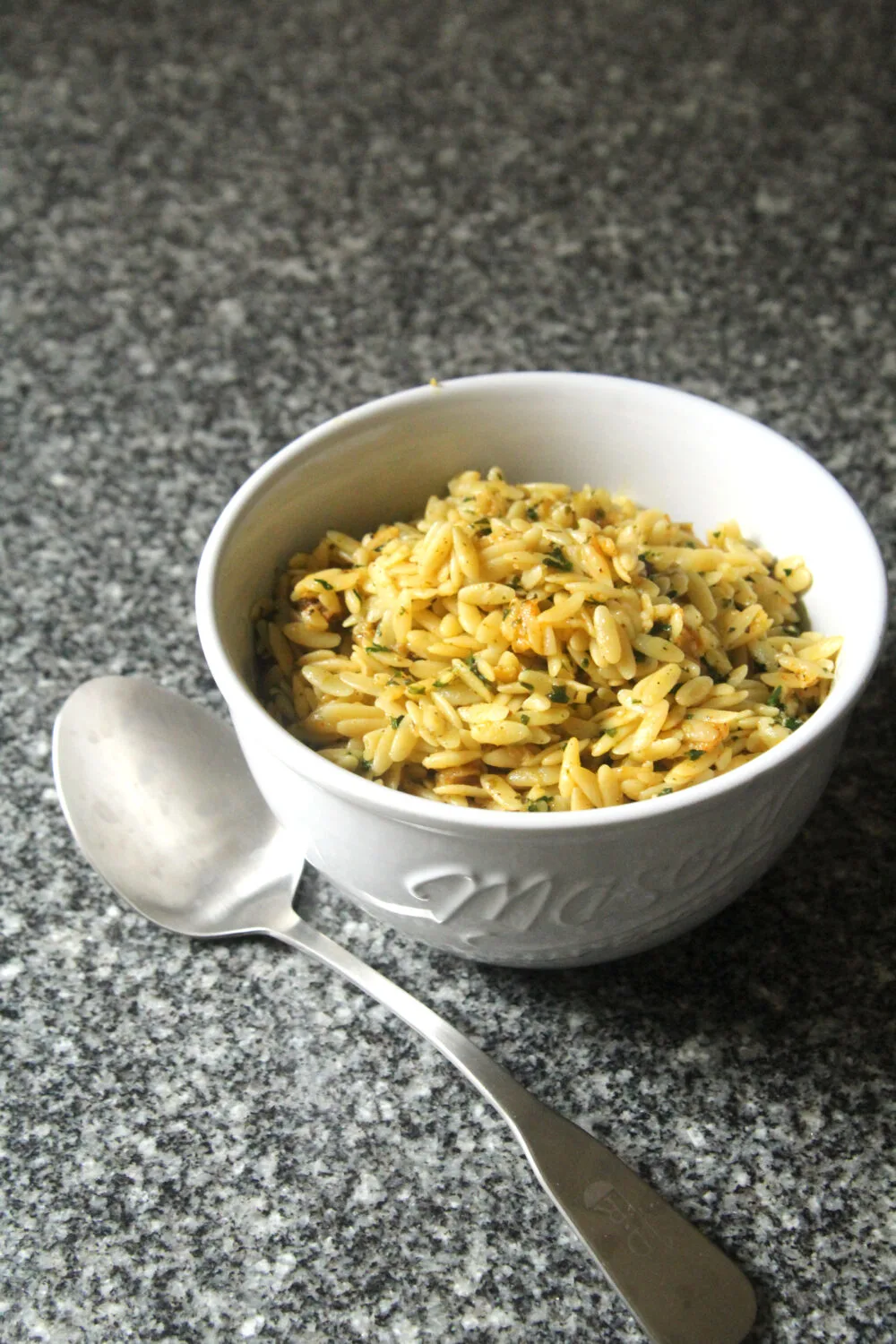 A white bowl holds yellow-ish lemon butter orzo on a granite countertop. A silver serving spoon sits nearby.