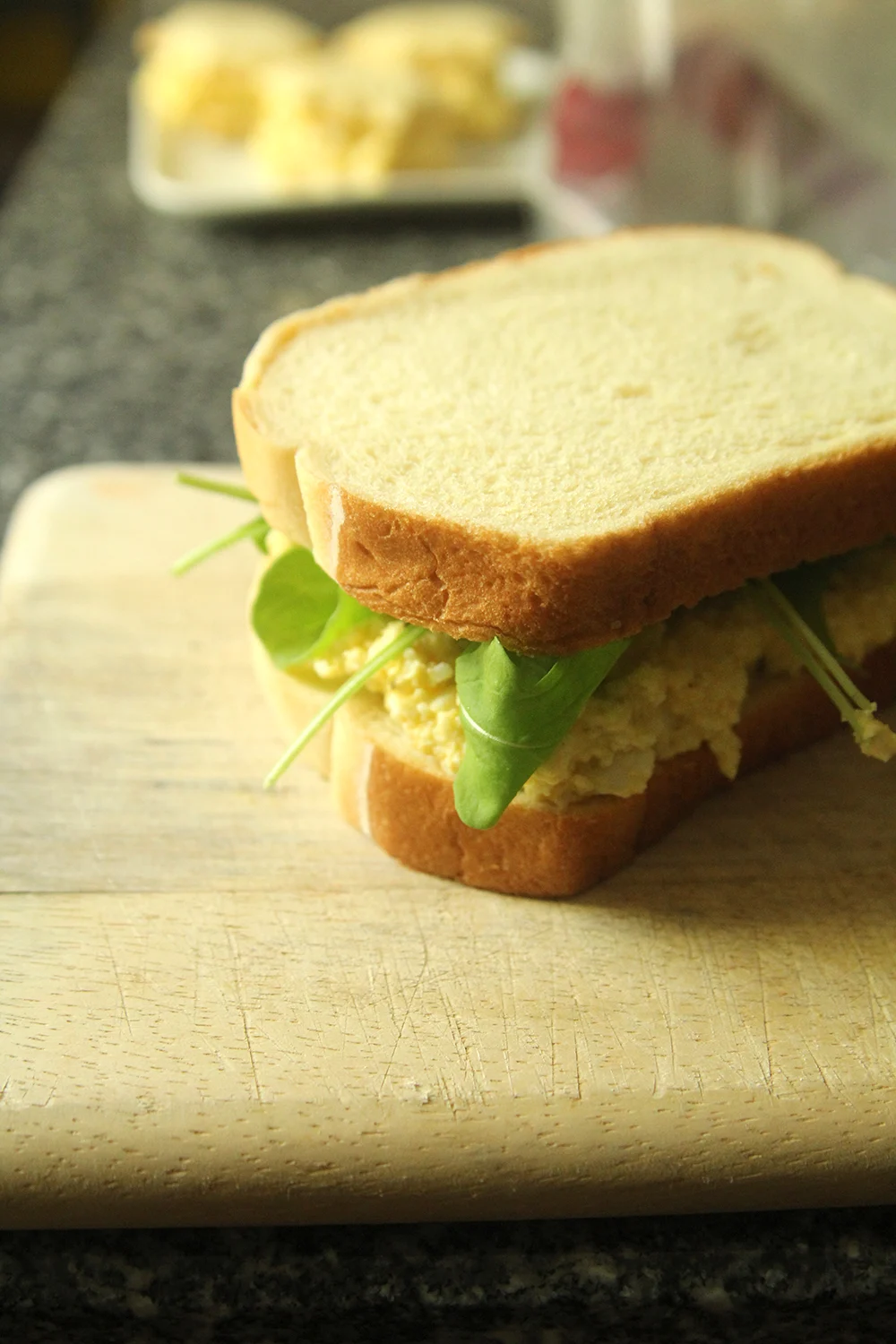A sandwich made with egg salad is shown on a cutting board. Green baby arugula pokes out in all directions.