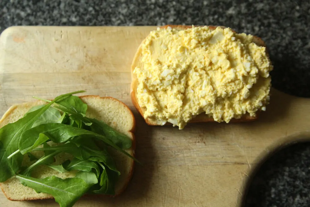 Two slices of bread sit on a wooden cutting board. One is topped with easy egg salad. The other is topped with green arugula.
