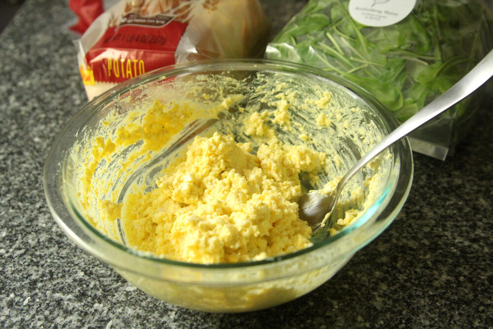 Easy egg salad is shown in a glass mixing bowl with a spoon inside. Nearby is a loaf of bread and a bag of arugula.
