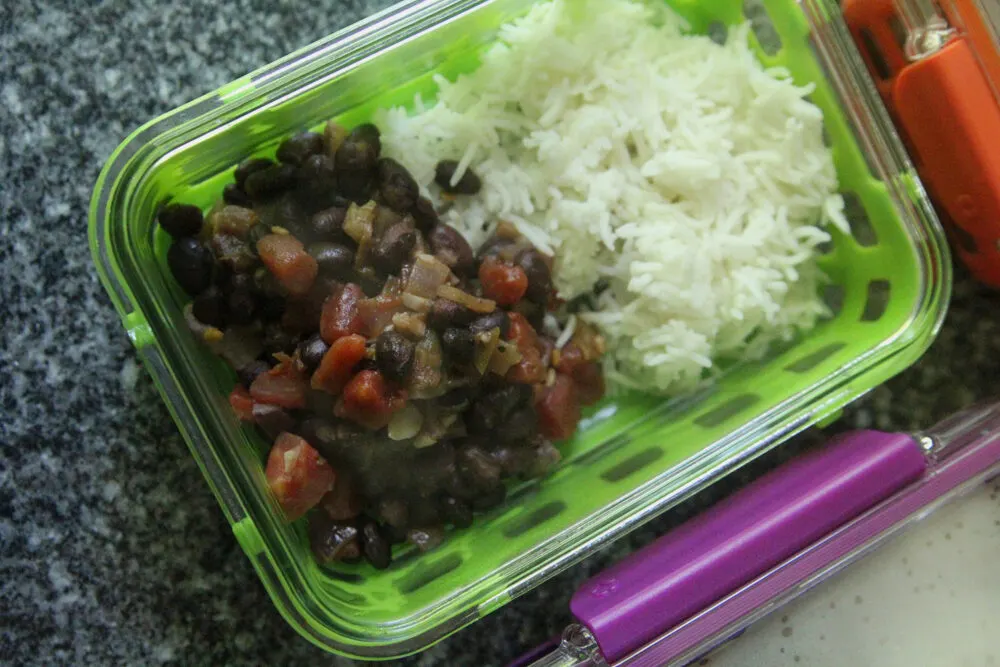 Steaming black beans with tomatoes and onions are shown with white rice in a green meal prep container.
