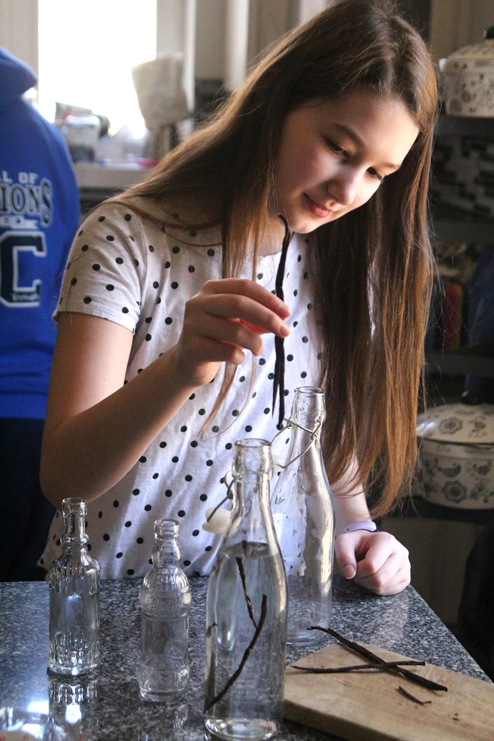 A young girl places vanilla beans into a glass jar filled with clear liquid. This photo is from Sarah's Cucina Bella. All rights reserved.