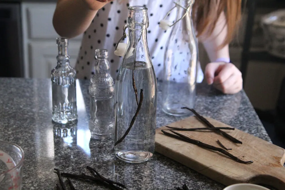 Vanilla beans are seen being placed into a bottle filled with clear liquid. Other vanilla beans sit nearby on the counter and on a cutting board.